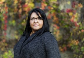 An Indigenous woman with long dark hair and glasses stands against a colourful fall backdrop.