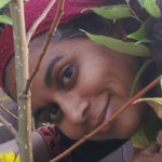 An Indian woman with brown skin and dark hair looks at the camera from an angel, surrounded by leafy green trees.