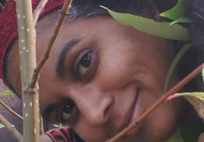 An Indian woman with brown skin and dark hair looks at the camera from an angel, surrounded by leafy green trees.