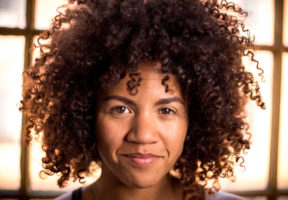 A young BLack woman with a large Afro stands against a bright window.