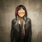 A middle-aged Indigenous woman in a black leather jacket. She is smiling and has red streaks in her long dark hair.