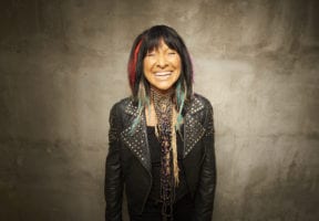 A middle-aged Indigenous woman in a black leather jacket. She is smiling and has red streaks in her long dark hair.