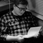 An Indigenous man wearing a checkered sweater and dark-rimmed glasses.