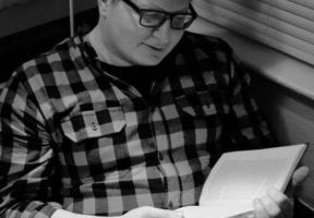 An Indigenous man wearing a checkered sweater and dark-rimmed glasses.