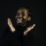 A young Black woman smiles and frames her face with her hands against a dark background.