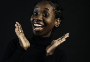 A young Black woman smiles and frames her face with her hands against a dark background.