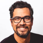 An Indigenous man with a goatee and dark-rimmed glasses smiels against a white background.