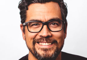 An Indigenous man with a goatee and dark-rimmed glasses smiels against a white background.