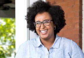 A photo of a Somali woman with curly hair. She wears dark-rimmed glasses and is laughing.