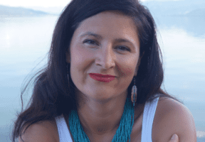 An Indigenous woman with long dark hair sits against a misty lake background.