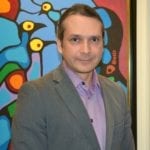 AN Indigenous man with hair pulled back in a ponytail. He wears a dark grey suitjacket and a purple collared shirt.