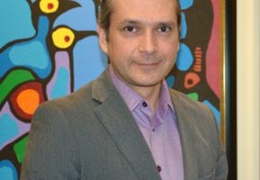 AN Indigenous man with hair pulled back in a ponytail. He wears a dark grey suitjacket and a purple collared shirt.