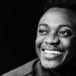 A black and white photo of a young Black man, smiling against a black background.