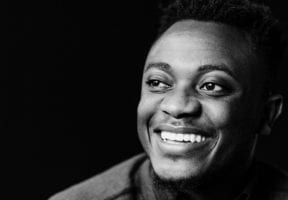 A black and white photo of a young Black man, smiling against a black background.