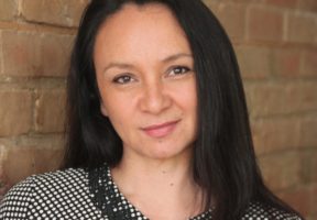 A white woman with long brown hair stands smiling against a brick wall.