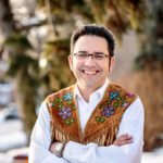 A Indigenous man wearing a white buttoned shirt and a brown beaded vest.