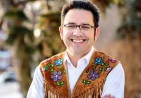 A Indigenous man wearing a white buttoned shirt and a brown beaded vest.