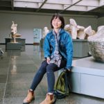 An Asian woman with a short bob sits in the middle of a gallery of art sculptures. She wears a blue denim jacket and blue jeans.