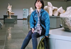 An Asian woman with a short bob sits in the middle of a gallery of art sculptures. She wears a blue denim jacket and blue jeans.