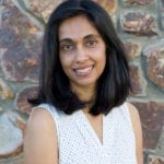 An Indian woman in a white tank top, standing against a brick wall.