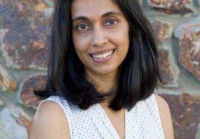 An Indian woman in a white tank top, standing against a brick wall.