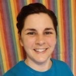 A white woman with brown hair pulled back in a ponytail smiles at the camera. She is wearing a blue t-shirt.