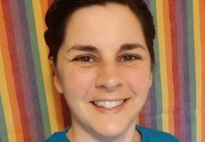 A white woman with brown hair pulled back in a ponytail smiles at the camera. She is wearing a blue t-shirt.