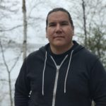 An Indigenous man with long dark hair in a ponytail. He stands against a bright winter tree background.