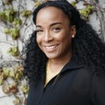 A young Black woman smiles against a leafy green background