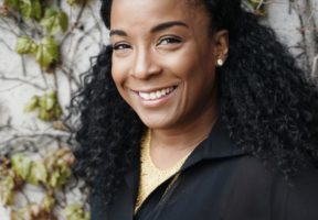 A young Black woman smiles against a leafy green background