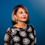 A photo of an older South Asian woman in front a dark blue background wearing a blakc blouse with a white pattern.