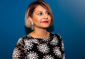 A photo of an older South Asian woman in front a dark blue background wearing a blakc blouse with a white pattern.