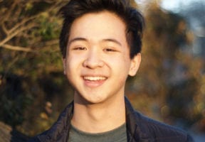 A young asian man outside wearing a grey t-shirt under a black quilted jacket