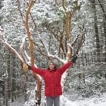 A faraway shot of a woman in a red winter jacket standing in winter jacket and a tree behind her