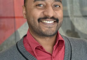 A south asian man with a red dress shirt under a grey and black cardigan