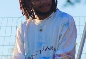 Headshot of William Padilla-Brown. He's a Black man with short dreads and wearing a white long sleeved shirt outside.