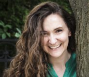 A young white woman stands beside a tree trunk and smiles. She wears a green button-down shift top and a cream cardigan. She has long curly brown hair.