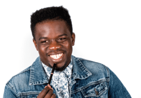 A Black man with cropped hair. He wears a denim jacket over a white skirt and is standing against a white background.