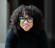 A Black woman wearing green-rimmed glasses smiles at the camera. She wears a black turtleneck top.