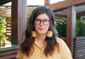 An Indigenous woman with long dark hair stands against a city courtyard background. She wears glasses and Indigenous beaded feather earrings.