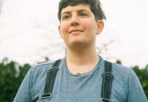 A white non-binary individual with close-croppsed brown hair looks off into the distance and smiles. They are wearing a blue t-shirt and overalls.