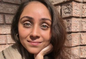 A young Indian woman with long brown hair and hazel eyes sits against a brick background.