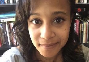 A young Black woman with shoulder-length dark hair and bangs. She wears a grey shirt and stands in front of a bookshelf.