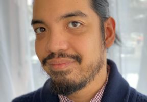 A dark-haired Filipino man with a moustache and scruffy beard smiles gently in front of a window.