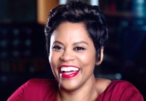 A Black woman in a deep red blouse smiles widely at the camera.
