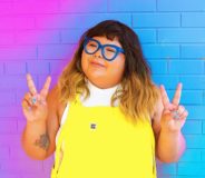 An Indigenous woman with long ombre hair that runs from brown to orange-blonde. She wears a pair of bright yellow overalls, blue-rimmed glasses, and is holding her hands up in two peace signs (index and middle fingers held aloft). She stands against a bright purple-blue background wall.