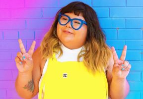 An Indigenous woman with long ombre hair that runs from brown to orange-blonde. She wears a pair of bright yellow overalls, blue-rimmed glasses, and is holding her hands up in two peace signs (index and middle fingers held aloft). She stands against a bright purple-blue background wall.