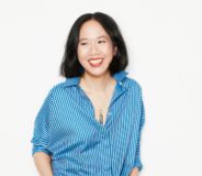 A Chinese-Canadian woman stands against a white background with her hands in her pockets. She has short dark hair and wears a blue denim button-down shirt over dark pants, and is smiling widely.
