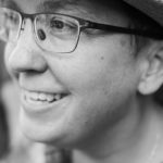 A black-and-white photo of a Latinx woman. She wears glasses and a hat with a brim.