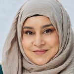 A woman with dark eyes and a beige hijab smiles at the camera.